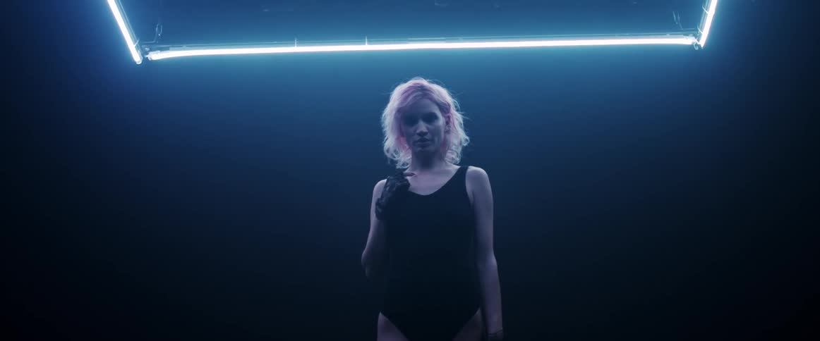A woman with short blond hair stands and poses in a darkened room illuminated by neon lights