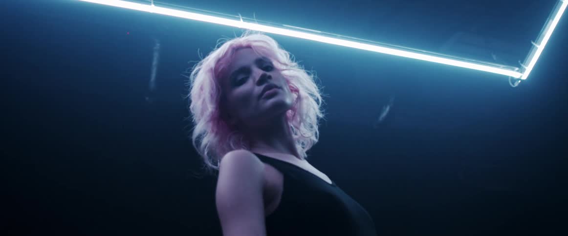 A woman with pink hair in a room with a white neon light strip staring intensely at the camera