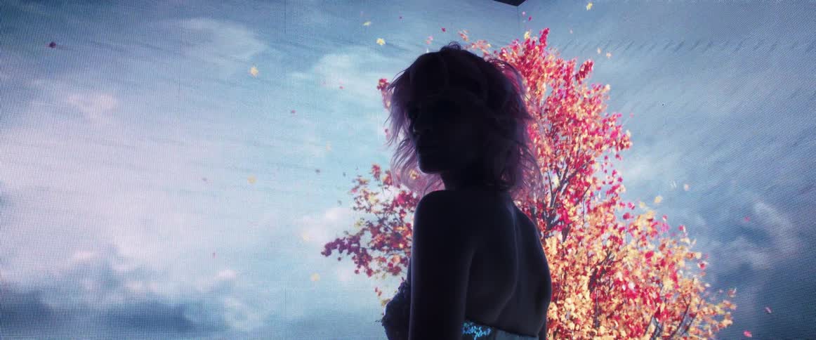A woman gazing into the camera turns away to face a video wall displaying a tree shedding red leaves