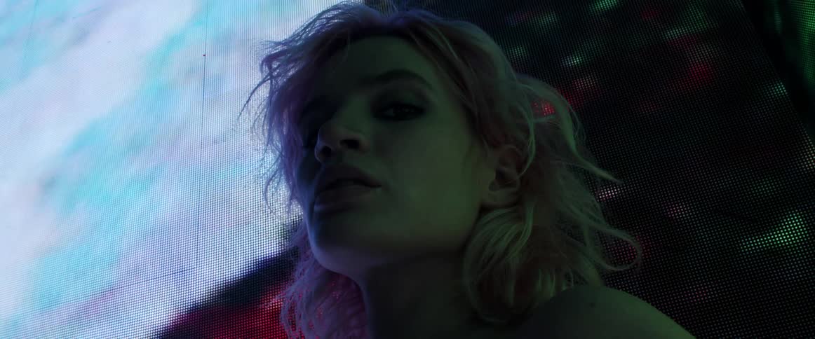 A woman with blonde hair is illuminated by a large digital screen displaying digital imagery behind her