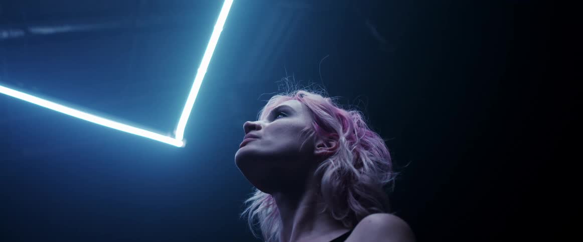 A woman with pink hair looking up in a dark room with a white neon light strip over her head