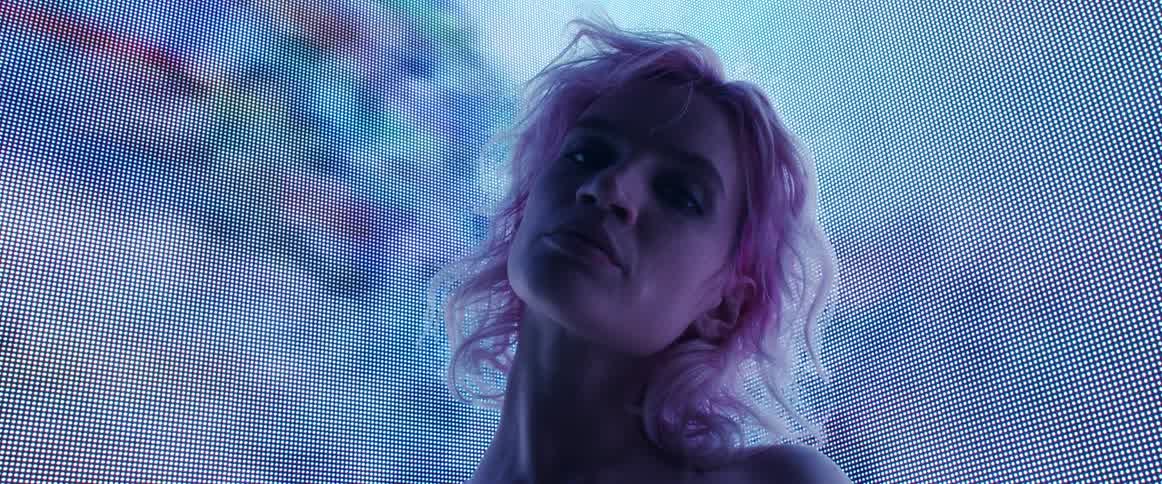 Young woman with pink hair looking at the camera in front of giant digital displays