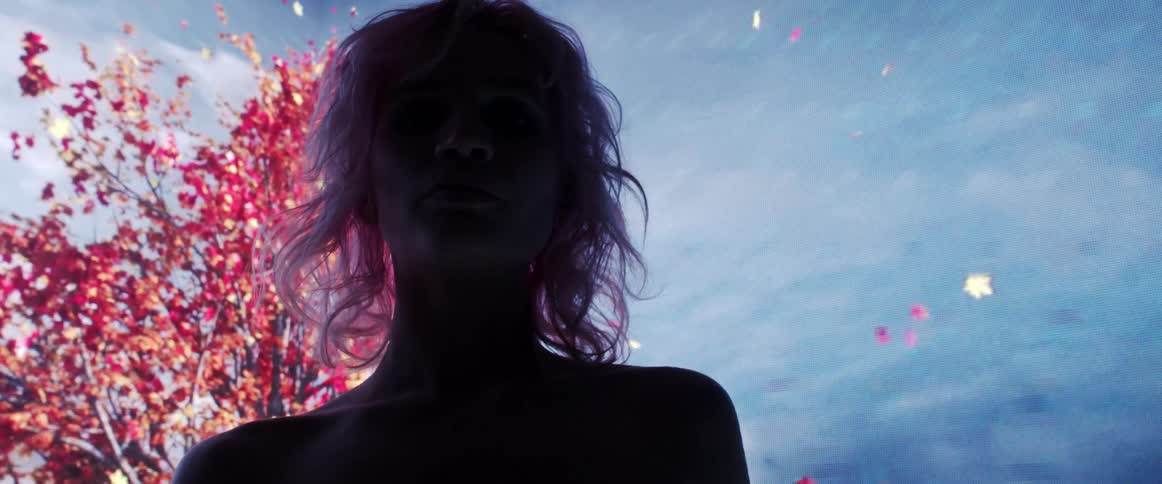 The silhouette of a woman is illuminated by a large screen displaying trees and falling leafs behind her