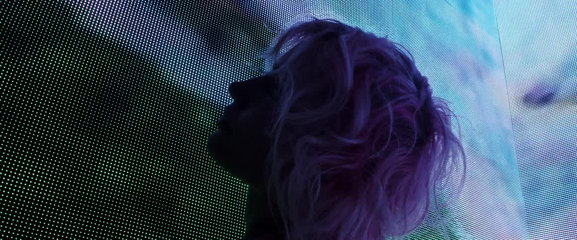 A woman gazes into the camera while standing in the corner of a video wall before turning her face away