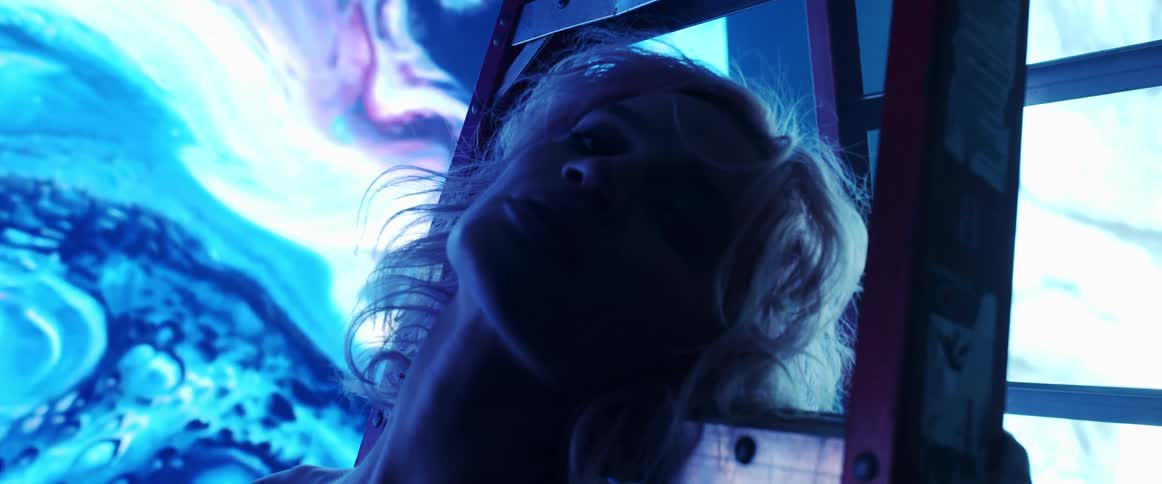 A woman with short blonde hair stands and is illuminated by a large digital screen displaying abstract imagery behind her