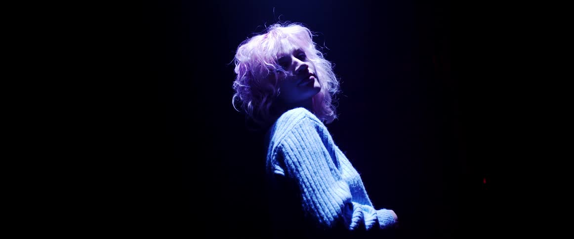 A woman with short blond hair stands and poses in a darkened room illuminated by neon lights