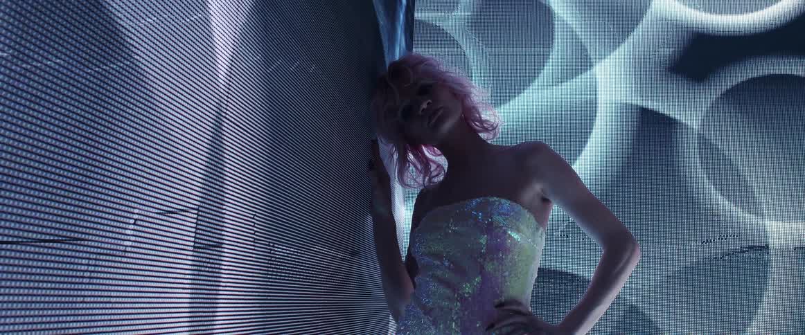 Young woman with pink hair looking at the camera on a dark room in front of a digital display