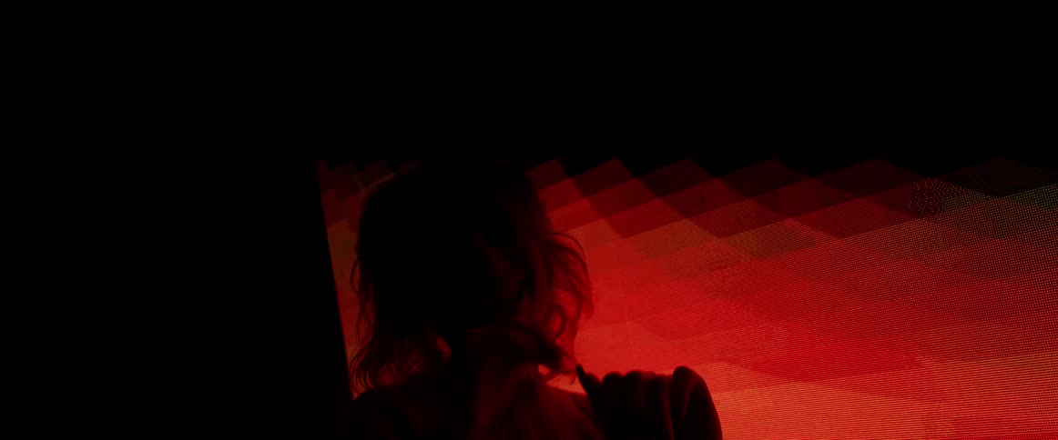 A woman with pink hair dances around playfully in a dark room in front of a digital screen changing colors