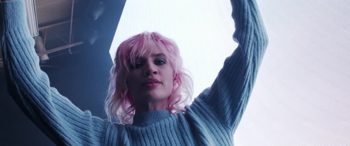 A young woman with blond hair looks at the camera while raising her arms up while dancing
