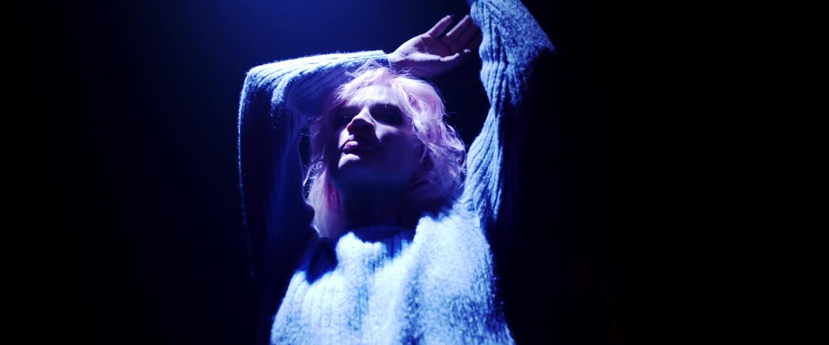 A woman with short blond wearing a sweater raises her arms beneath a soft neon light in a dark room