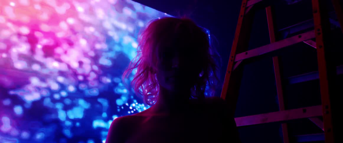 A woman with short blond hair stands in front of a huge screen displaying beautiful abstract imagery