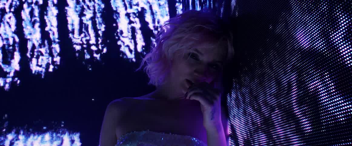 A woman standing in a room with black and purple digital screens shifting images all around her posing and staring at the camera
