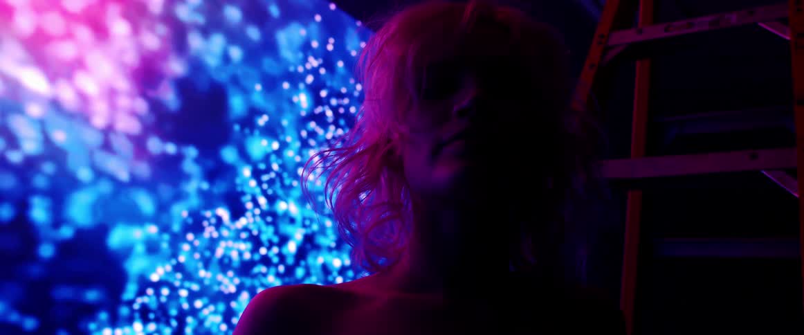 The silhouette of a woman with short blonde hair is illuminated by a large digital screen displaying digital imagery behind her