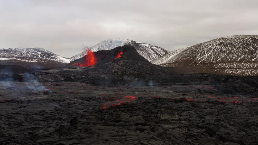 Erupting volcano spews molten hot lava into the air with snow covered mountains in the distance