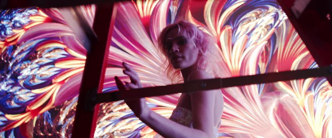 A woman with short blond hair walks in front of a large screen with colorful digital imagery displayed on it