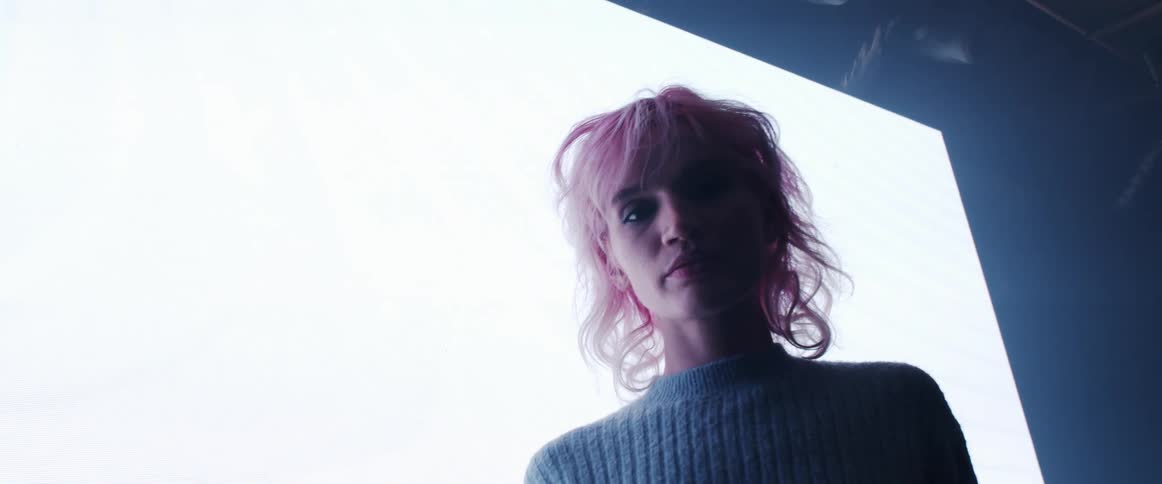 Young woman with pink hair looking at the camera on a dark room in front of a digital display