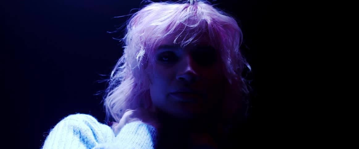 A woman with short blond wearing a sweater poses while illuminated by a soft violet neon light while in a darkened room