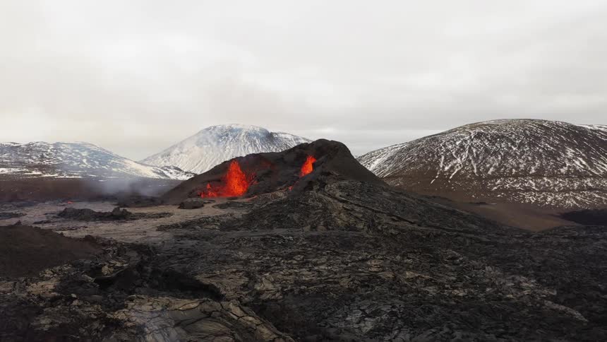 A volcano spewing molten lava with smoke with a mountain range the sky and clouds in the background