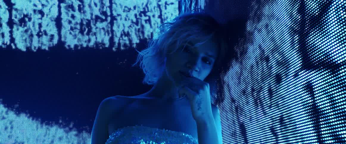 A woman with short blond hair leans against a huge screen displaying digital imagery