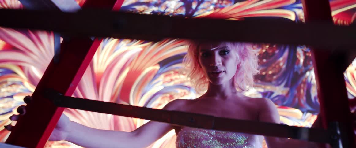 A woman walks behind a red structure and walks backwards while staring at the camera in front of a moving art backdrop