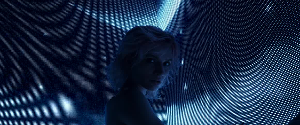 A woman stands in a darkened room in front of a large screen displaying digital imagery