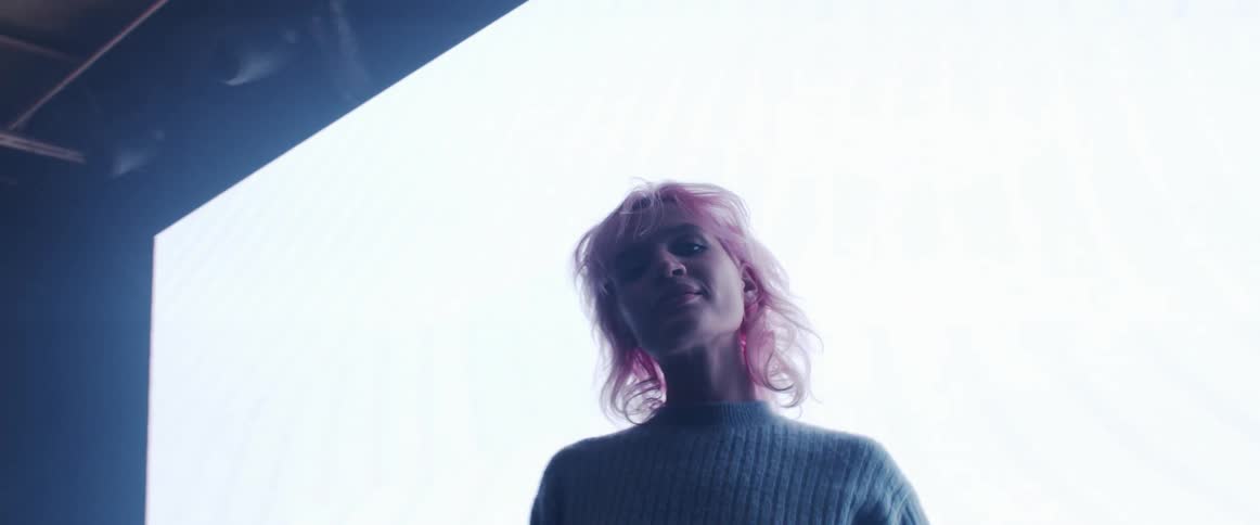 A young woman gazes into the camera while standing in front of a video wall in a dark room