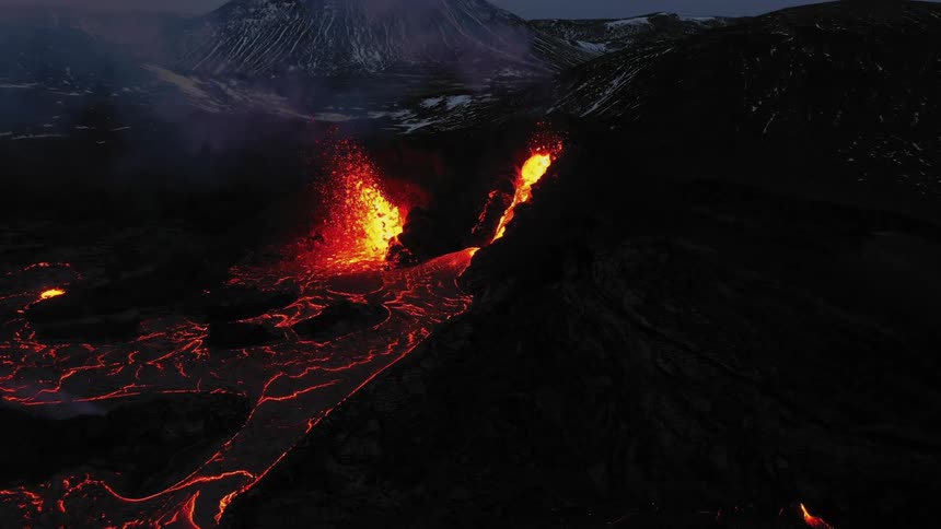 Erupting volcano spews molten hot lava into the air and oozes a river of hot lava with snow covered mountains in the distance