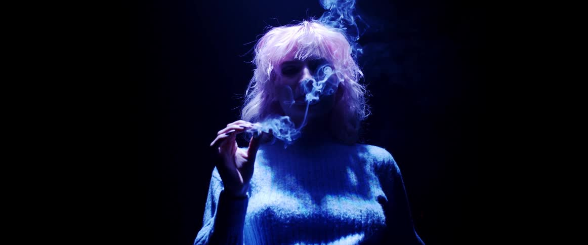 A woman with short blond hair stand under a soft neon light while smoking in a darkened room