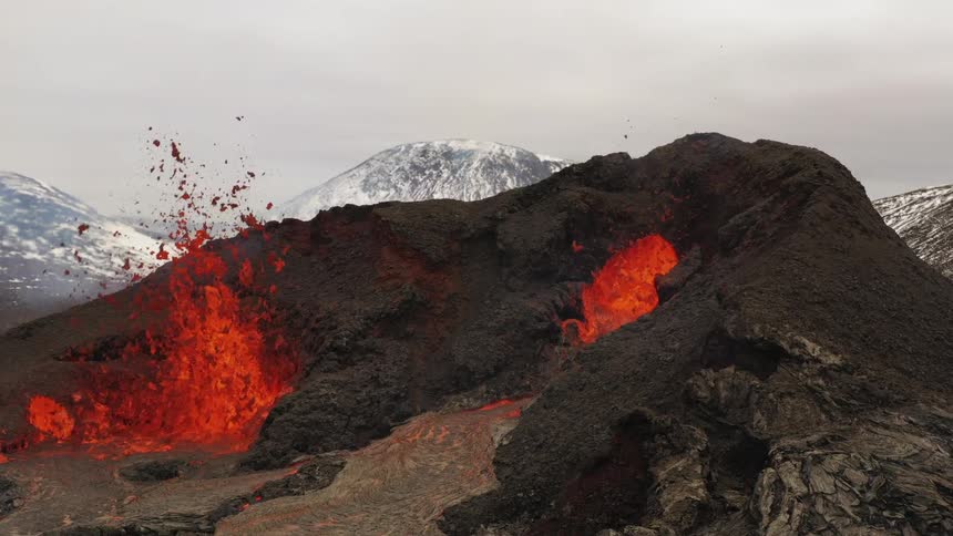 Erupting volcano spews molten hot lava into the air and oozes a river of hot lava with snow covered mountains in the distance