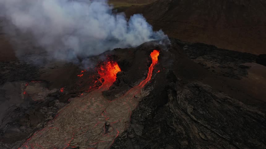 Smoke rises as an erupting volcano spews molten hot lava and oozes a river of hot lava