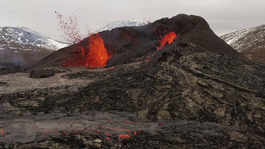 Erupting volcanic crater spews molten hot lava into the air with snow covered mountains in the distance