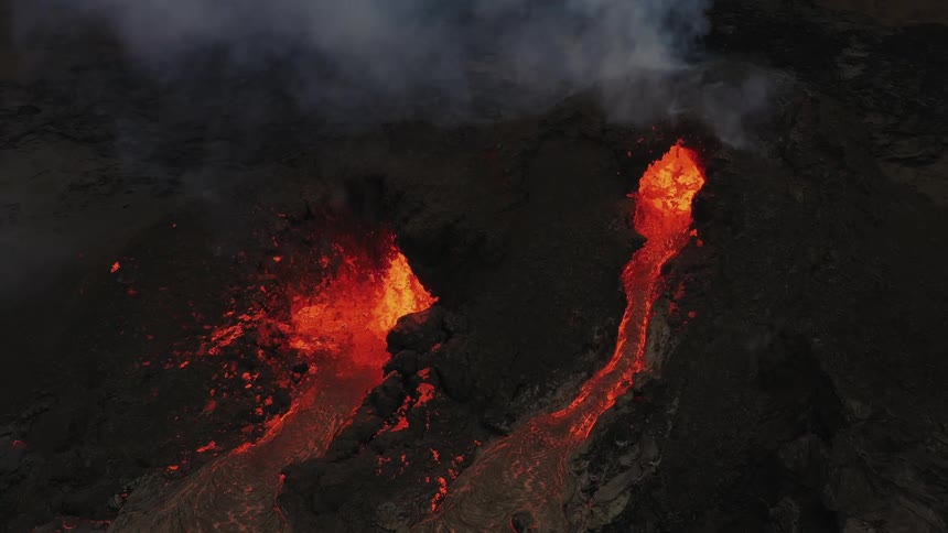 Smoke rises as an erupting volcano spews molten hot lava and oozes a river of hot lava