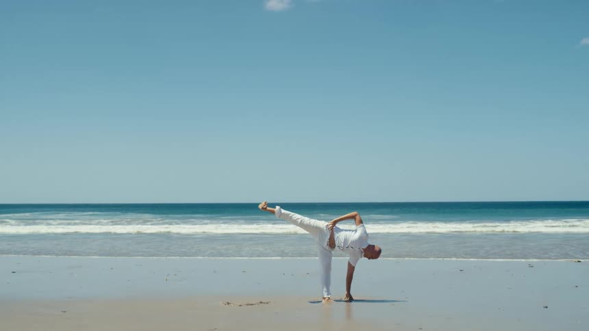 Man balances on hand and leg in yoga pose on beach on windy day