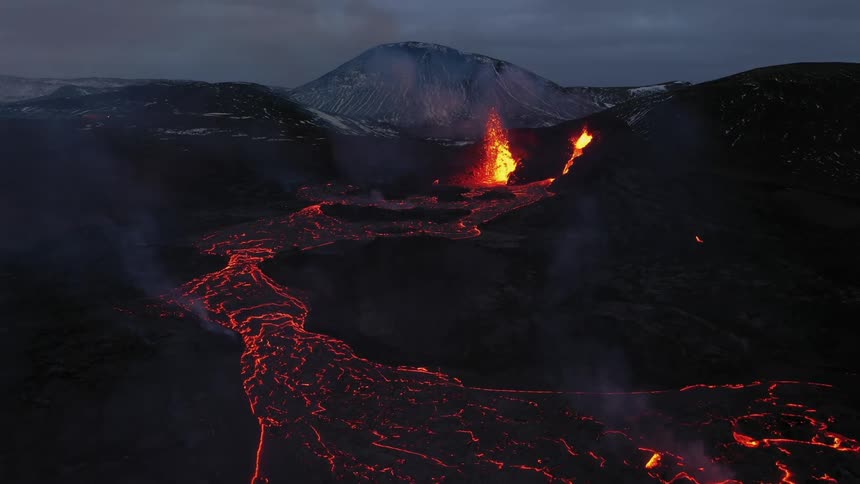 Erupting volcano spews molten hot lava into the air and oozes a river of hot lava with snow covered mountains in the distance