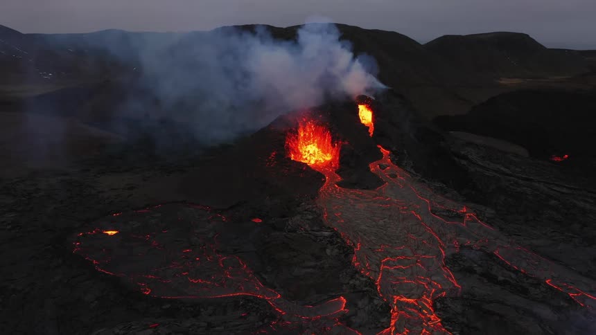 Smoke rises as an erupting volcano spews molten hot lava and oozes a river of hot lava