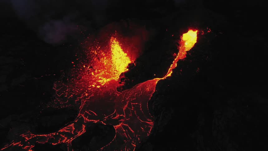 Erupting volcano spews molten hot lava into the air as hot lava flows and glows in the darkness