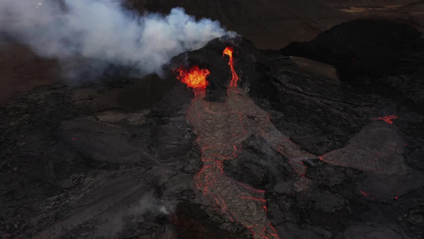 Smoke rises as an erupting volcano spews molten hot lava and oozes a river of hot lava