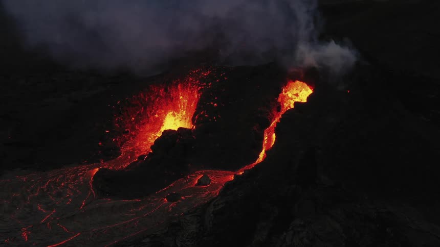 Erupting volcano spews molten hot lava into the air as hot lava flows and glows in the darkness