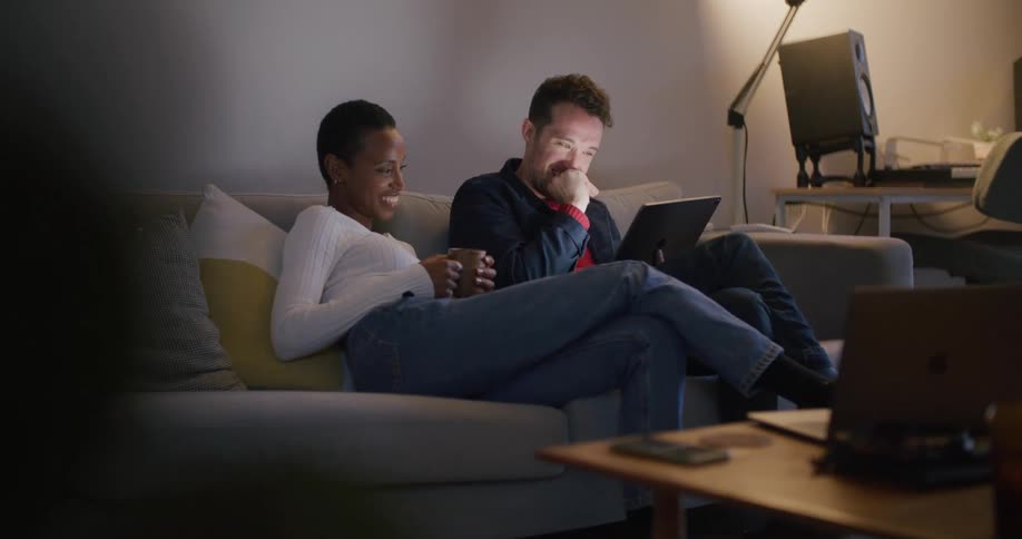 A man and woman sit together on a couch laughing at a tablet screen