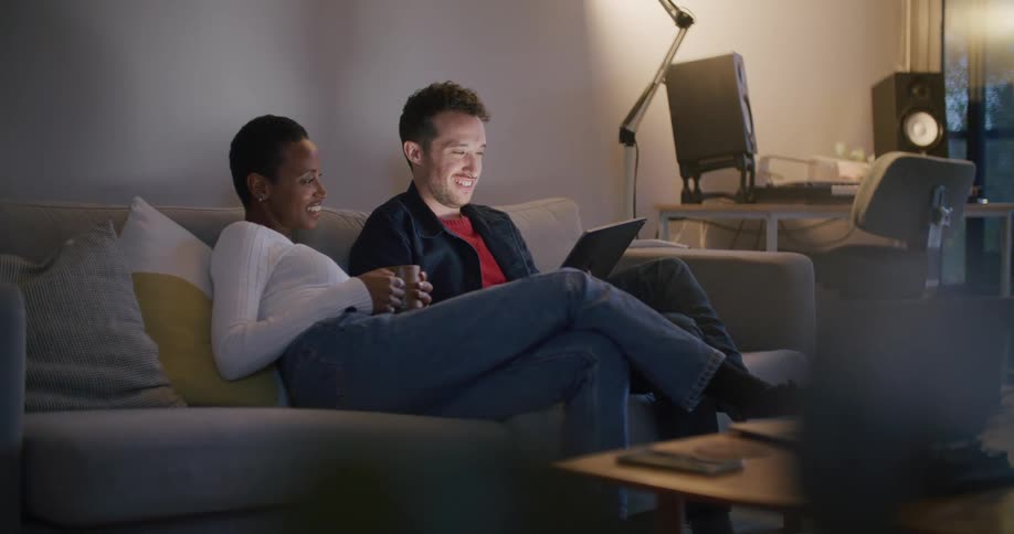 A man and woman laugh as they sit on a couch as they look at a tablet together