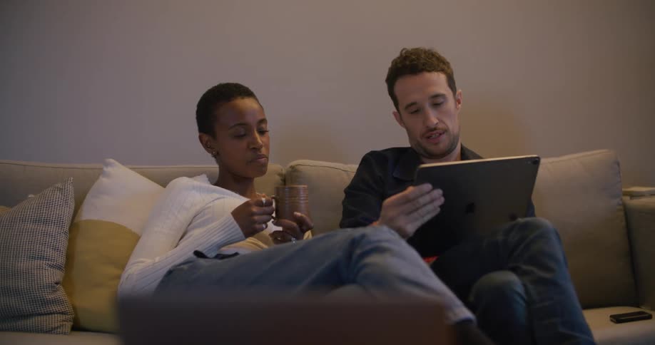 Man and woman sitting on a couch drink coffee and use a tablet in the living room at home