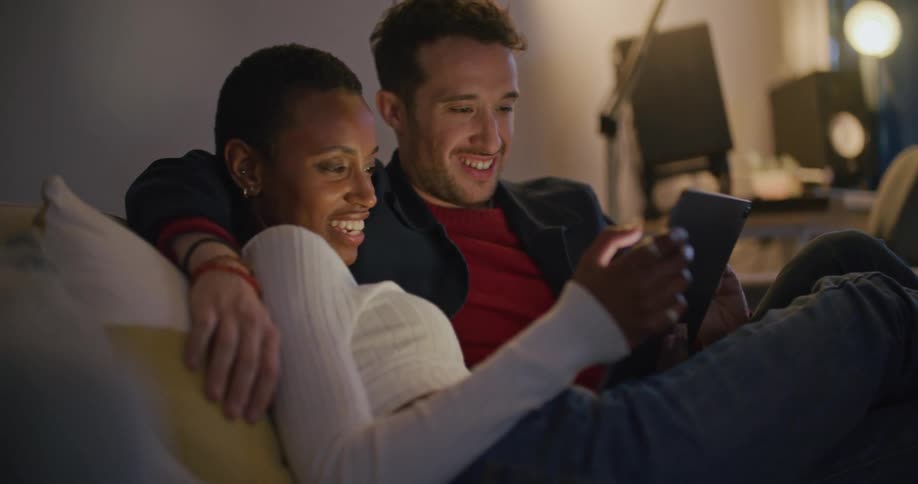 A man and woman watch a tablet while sitting together on a sofa indoors
