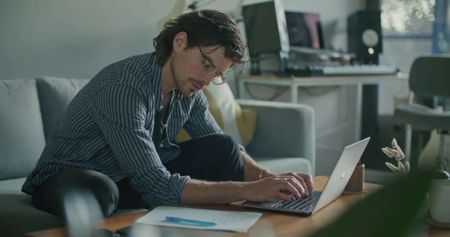 A man typing on a laptop stops to write on a sheet of paper while sitting on a sofa in the living room of a house during the day