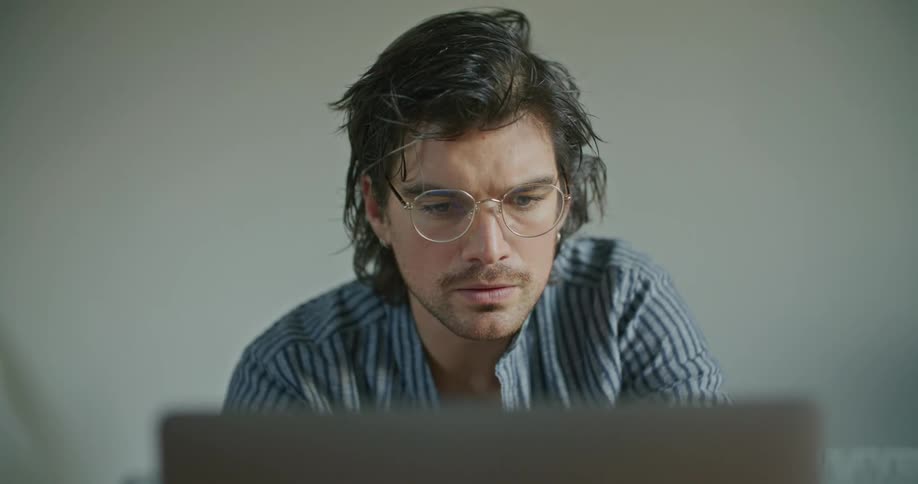 A man with glasses in a room looking at a laptop with a wall in the background