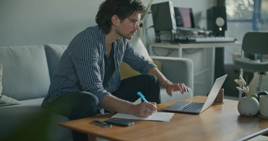 A man sits on a couch writing on a piece of paper that is on a table in front of him then looks at a laptop