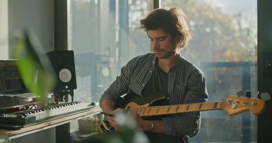 A man sits at home and holds a guitar while he uses his cell phone