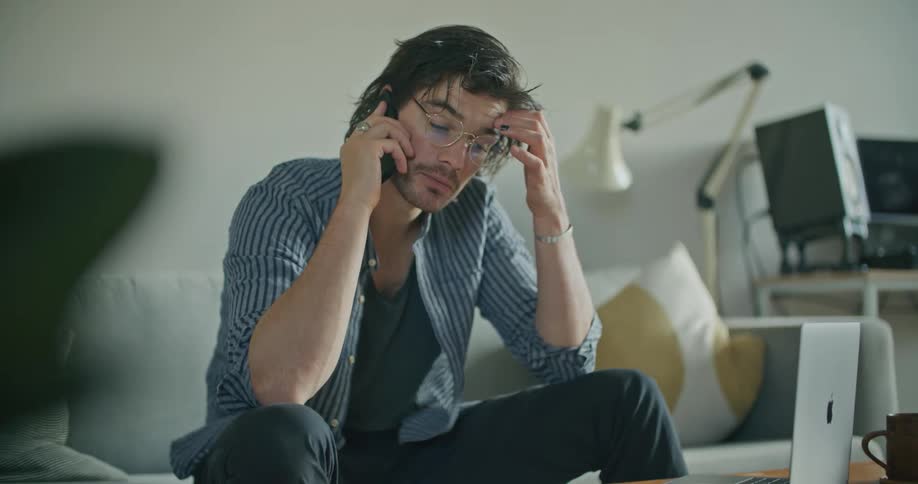A man in a room sitting on a couch talking on a smart phone and looking at a laptop