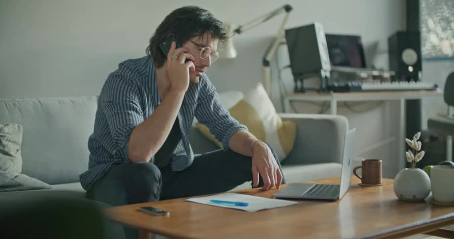 A man talks on a smartphone while looking at a laptop as he sits on a sofa in the living room of a house during the day