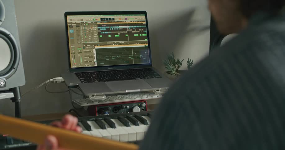 A man in a room playing a guitar with a keyboard and a laptop in front of him