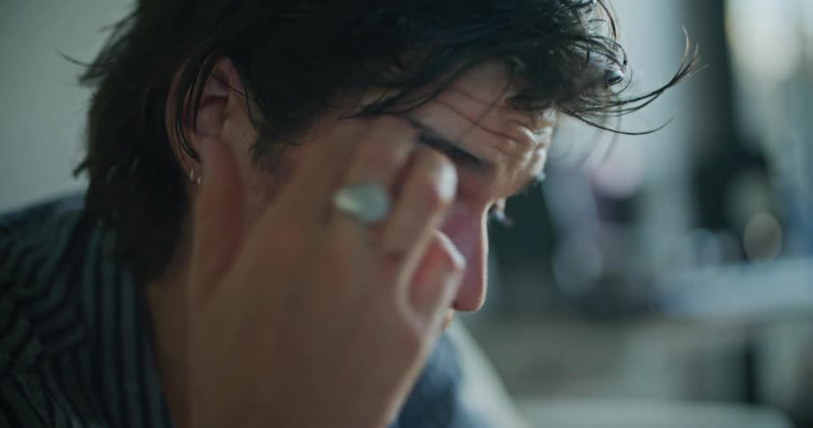 A man sits at home and takes his glasses off and rubs his eyes and puts his glasses back on